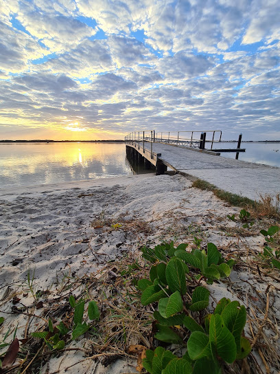 A serene lakeside scene during sunset with a wooden pier extending over calm water. The sky is partly cloudy, reflecting warm hues over the lake. In the foreground, there's sandy shore with green plants growing near the water's edge.