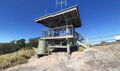 A small, elevated lookout tower with railings and antennas is situated on a rocky hilltop under a clear blue sky. The structure has a metal roof supported by wooden beams. The surrounding landscape includes grassy patches and distant trees.