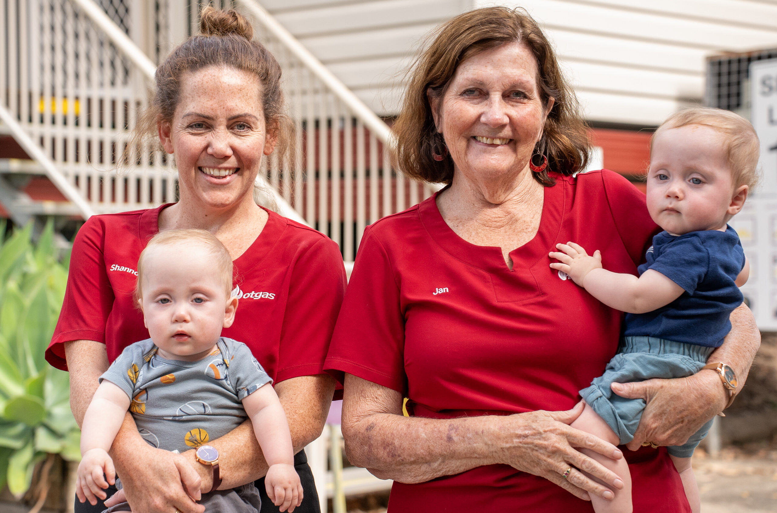 Two women in red shirts each hold a baby. They are standing in an outdoor setting with a staircase and building in the background. The woman on the left has a name tag that reads "Shannon," and the woman on the right has a name tag that reads "Jan.